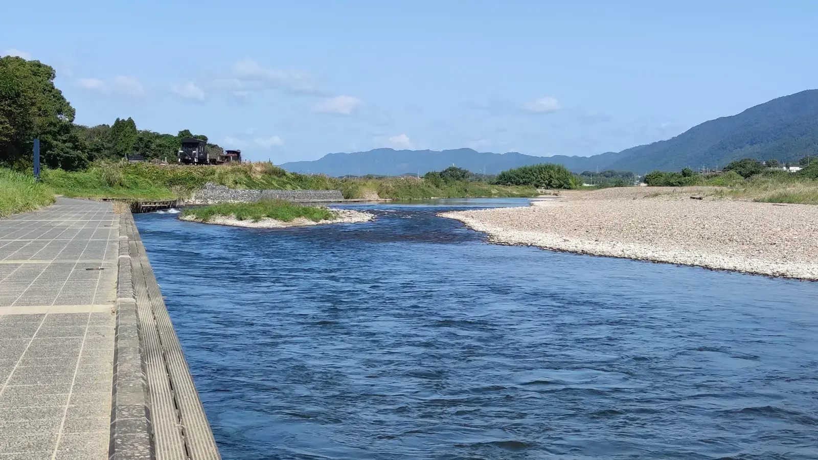 亀岡市を流れる保津川。手前に川沿いの遊歩道、川の向こうに白い河原と山並みが続き、青空が広がっている